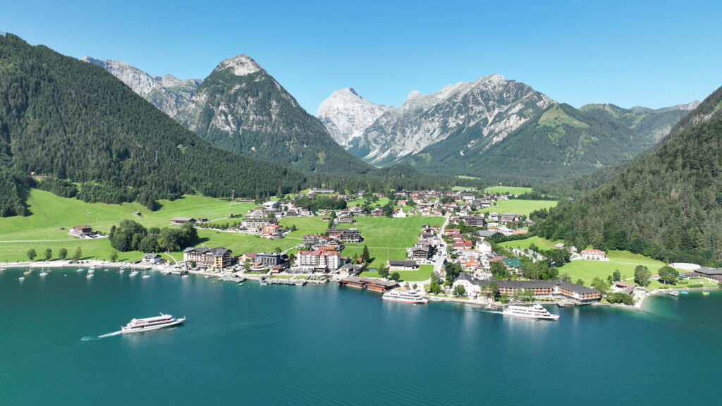 Der Blick schweift hinunter auf die charmante Ortschaft Pertisau am Achensee. Entlang des Seeufers legen zahlreiche Segelboote an, während die Schiffe der Achenseeschifffahrt ihre Runden drehen.
